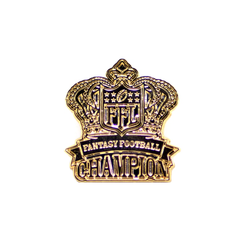 Op de afbeelding: Een goudkleurige Fantasy Football Champion-speld. De speld heeft een kroonontwerp met de tekst "FFL" en "Fantasy Football Champion" in zwarte letters. De speld staat tegen een witte achtergrond.