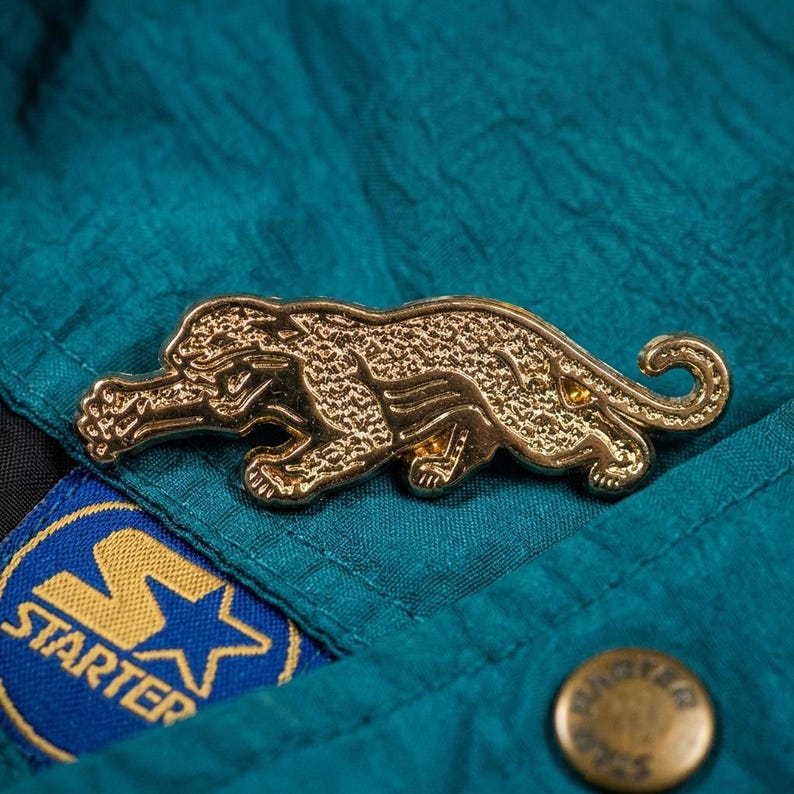 May include: A gold-colored metal pin in the shape of a jaguar, lying on a teal jacket. The pin is detailed with textured fur and is positioned above a Starter brand patch.