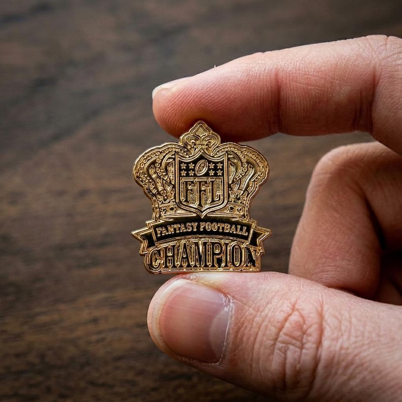 Op de afbeelding: Een goudkleurige Fantasy Football Champion-speld. De speld heeft de vorm van een kroon met de letters "FFL" en de woorden "Fantasy Football Champion" in zwarte letters. De speld wordt tussen twee vingers gehouden.