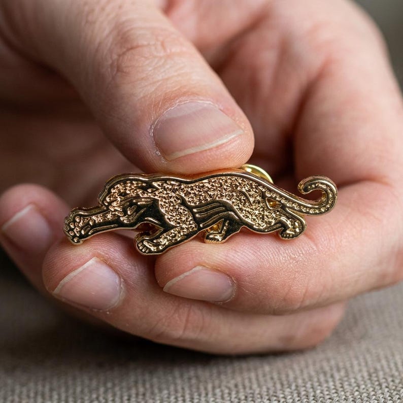 May include: A gold-colored pin in the shape of a jaguar. The pin is detailed with a textured pattern and is held in a person's hand. The jaguar is depicted in a crouching position, with its tail curled.