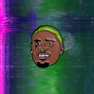 May include: A green and black enamel pin featuring a portrait of a person with a green mohawk, a black eye patch, and a gold nose ring. The person is smiling and has gold teeth.