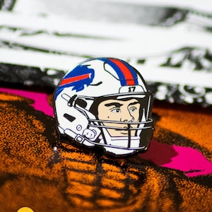May include: A white football helmet with a blue and red stripe, a blue buffalo logo, and the number 17 on the side. The helmet is a pin with a black and white cartoon face inside.