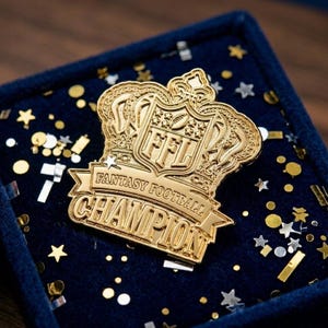 Op de afbeelding: Een goudkleurige Fantasy Football Champion-speld in een marineblauwe fluwelen doos. De speld heeft een kroonontwerp met de letters "FFL" en de woorden "Fantasy Football Champion". Confetti omringt de speld.