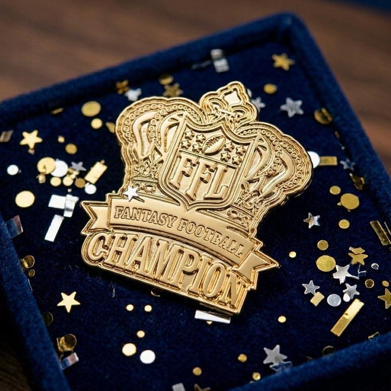 Op de afbeelding: Een goudkleurige Fantasy Football Champion-speld in een marineblauwe fluwelen doos. De speld heeft een kroonontwerp met de letters "FFL" en de woorden "Fantasy Football Champion". Confetti omringt de speld.