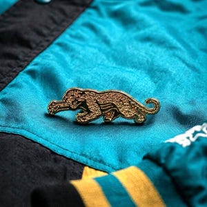 May include: A gold-colored pin in the shape of a jaguar, lying on a teal and black fabric. The pin is detailed with textured surfaces, and the jaguar is in a crouching position. The background includes a teal, black, and yellow striped fabric.