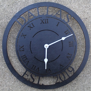 May include: Black wooden clock with Roman numeral markings and the words "Dallas Est 2019" around the edge.