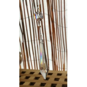 May include: A clear crystal prism with a faceted design hangs from a thin wire. The prism reflects a rainbow of colors. A smaller, round crystal is at the top. The background features vertical brown bamboo.