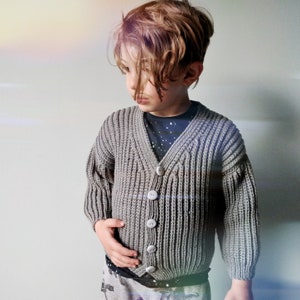 Puede incluir: Una persona joven lleva un cárdigan de punto gris con botones. El cárdigan tiene una textura acanalada y cuello en V.