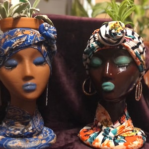 May include: Two decorative head planters with closed eyes, adorned with colorful head wraps. One is light-skinned with blue lips and eyeshadow, the other is dark-skinned with turquoise lips and eyeshadow. Each holds a small terracotta pot with greenery.