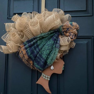 Pode incluir: Uma grinalda decorativa com a silhueta do rosto de uma mulher, adornada com um lenço xadrez verde e azul e um design de cabelo em malha bege. A mulher usa um colar e brincos de prata. A grinalda está pendurada numa porta azul escura.