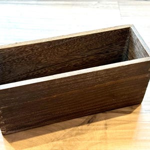 May include: A rectangular wooden box with a dark brown finish. The box is constructed from planks of wood, with visible grain and a natural, rustic appearance. The box is empty and has a simple, clean design.