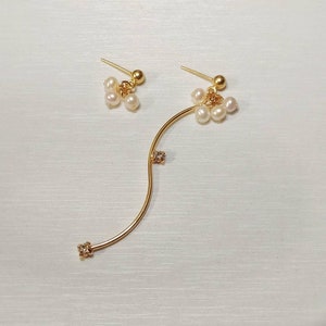 May include: A pair of gold-colored earrings. One is a stud with a cluster of small, white pearls. The other is a long, curved earring with a pearl cluster and three small, clear gemstones. The earrings are on a white surface.