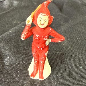 May include: A small figurine of a character wearing a red, pointed hood and a matching red bodysuit with white paint wear. The figure has a pale face, red lips, and is posed with one hand raised near the head and the other resting on the hip.