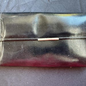 Puede incluir: Una cartera rectangular negra con un patrón texturizado de piel de lagarto. La cartera cuenta con un cierre de solapa horizontal asegurado con una fina barra metálica de tono plateado centrada a lo largo del borde de la solapa, diseño funcional.