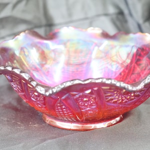 May include: A round carnival glass bowl featuring a vibrant red hue with an iridescent, rainbow-like sheen. The bowl has a scalloped, ruffled edge and a detailed pressed pattern consisting of geometric starbursts and vertical ribbed panels.