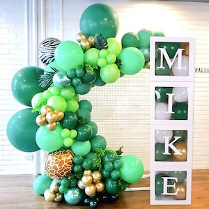 Puede incluir: Arco de globos verde, dorado y blanco con una caja blanca con las letras "MIKE" en negro. Los globos est&aacute;n dispuestos en forma de semic&iacute;rculo.