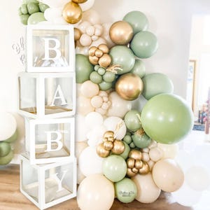 May include: A stack of four clear acrylic boxes with white frames spelling out the word "BABY". The boxes are decorated with green and gold balloons.