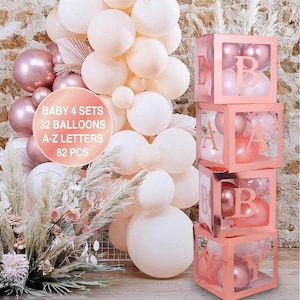 Puede incluir: Cuatro cajas transparentes apiladas con marcos de color oro rosa y letras que deletrean "BABY". Cada caja contiene globos en tonos de rosa y blanco. Las cajas est&aacute;n frente a una pared de globos en tonos de blanco y rosa.