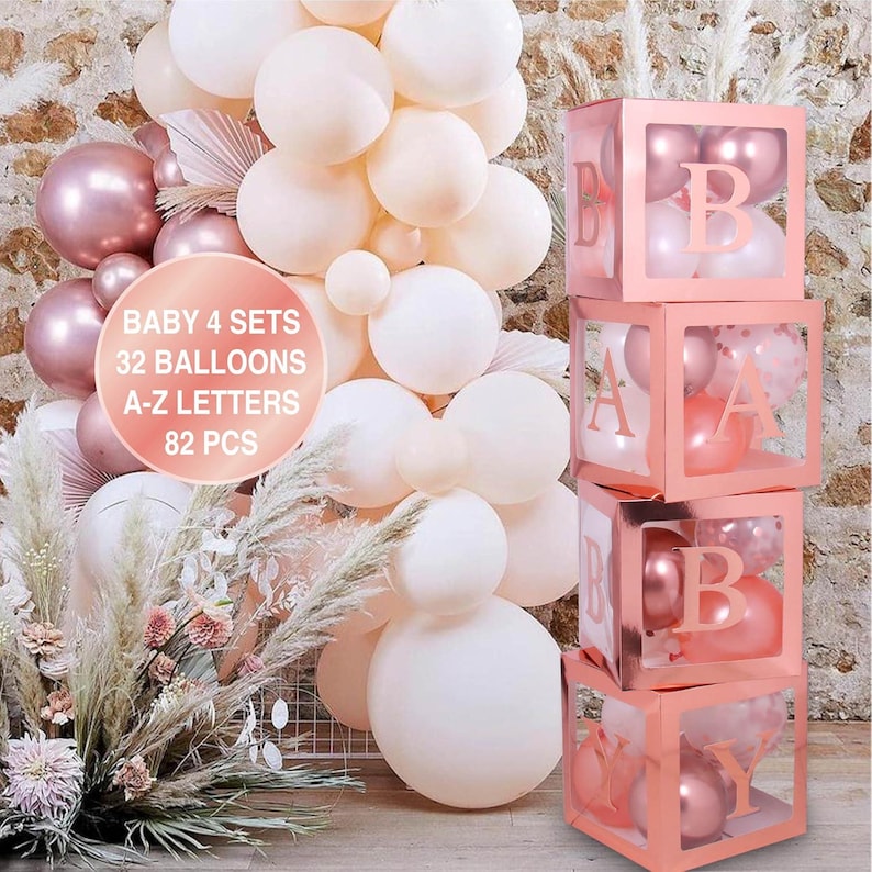 Puede incluir: Cuatro cajas transparentes apiladas con marcos de color oro rosa y letras que deletrean "BABY". Cada caja contiene globos en tonos de rosa y blanco. Las cajas est&aacute;n frente a una pared de globos en tonos de blanco y rosa.