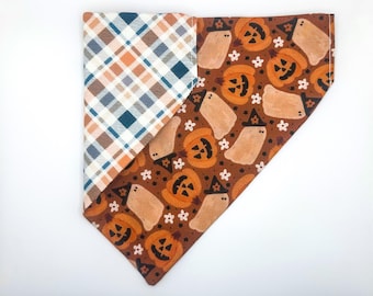 May include: Reversible dog bandana with a plaid pattern in blue, orange, and cream on one side. The other side features a Halloween-themed print with orange jack-o'-lanterns, ghosts, and white flowers on a brown background.