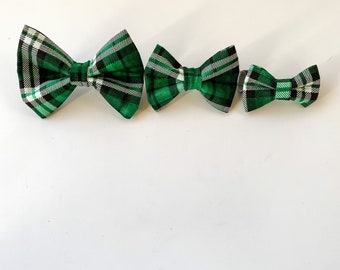 May include: Three green plaid bow ties of varying sizes are arranged on a white surface. The bow ties feature a pattern of green, black, and white stripes. The bow ties are likely for pets or accessories.