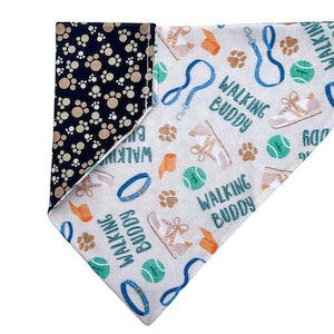 Puede incluir: Un bandana beige para perro con un patrón azul, verde y naranja. El bandana presenta el texto "Walking Buddy" e imágenes de pelotas de tenis, correas para perros y zapatos para perros. El reverso del bandana es azul marino con huellas de patas marrones.