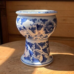 May include: A white ceramic pedestal bowl featuring a blue floral and vine pattern. The bowl has a wide, rounded upper section that tapers down to a narrower, flared base. The surface is decorated with intricate blue painted botanical motifs.