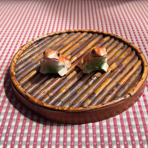May include: Two small ceramic frog figurines with orange and green coloring are positioned on a round, slatted bamboo tray. The frogs have dark eyes and are facing forward, resting on the dark brown, textured surface of the circular tray.