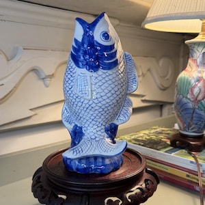 May include: A blue and white ceramic fish figurine featuring a detailed scale pattern and a vertical orientation, displayed on a dark brown carved wooden circular stand with four curved legs, showcasing a glossy finish and intricate painted details.