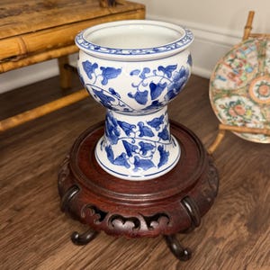 Può includere: Un vaso in porcellana bianca con un motivo blu di fiori e viticci, caratterizzato da una ciotola arrotondata e una base a piedistallo svasata. Il vaso è esposto su un supporto in legno intagliato marrone scuro con gambe curve e dettagli intagliati.