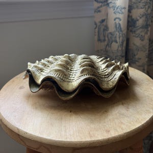 May include: A metallic gold-toned clam shell object with a textured, ribbed surface and a scalloped edge. The shell features a detailed, repeating pattern across its exterior, showcasing a curved, organic shape with a slightly weathered finish.