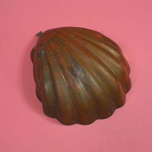 May include: A scalloped shell-shaped mold made of weathered copper with a dark, oxidized brown patina and visible streaks of green verdigris. The object features a series of deep, radiating ridges and a small metal hanging loop at the top edge.
