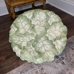 May include: A round ceramic serving platter shaped like a cluster of overlapping cabbage leaves. The dish features a cream-colored base with hand-painted sage green highlights along the embossed veins and ruffled edges of the individual leaves.