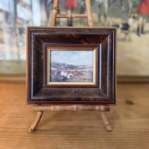 May include: A small rectangular painting featuring a fox hunting scene with riders on horses, encased in a thick, dark brown wood-grain frame with a thin gold inner border, displayed on a light brown bamboo easel stand.