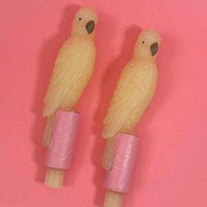 May include: Two identical parrot-shaped candles, each featuring a pale yellow wax bird perched on a textured pink cylindrical base. The birds have small black eyes and dark gray beaks, with detailed feather patterns molded into the wax surface.