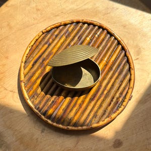 May include: A small, round brass container with a lid resting on a circular bamboo tray. The container has a smooth, metallic finish, while the tray is constructed from parallel bamboo stalks arranged in a circular frame with a warm brown tone.
