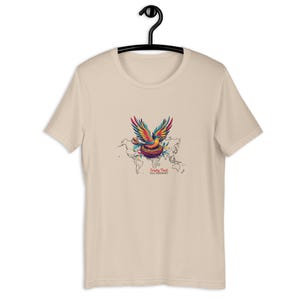 Puede incluir: Camiseta beige con un gráfico colorido de un pájaro con las alas extendidas sobre un nido y un mapa del mundo. El texto "Empty Nest, Full Passport" está debajo. La camiseta está colgada de una percha negra.