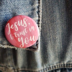 Könnte beinhalten: Rosa Knopf mit dem Text "Jesus, ich vertraue auf dich" in weißer Kursivschrift.