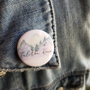 Puede incluir: Un botón blanco con un diseño de acuarela de montañas y árboles. El texto "Talitha Koun" está escrito en cursiva en el botón.