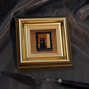 May include: A small, square painting in a gold frame depicts a black cat in a window. The cat is the focal point, with a white object in its mouth. The painting is set against a dark, textured background. A silver palette knife rests below.
