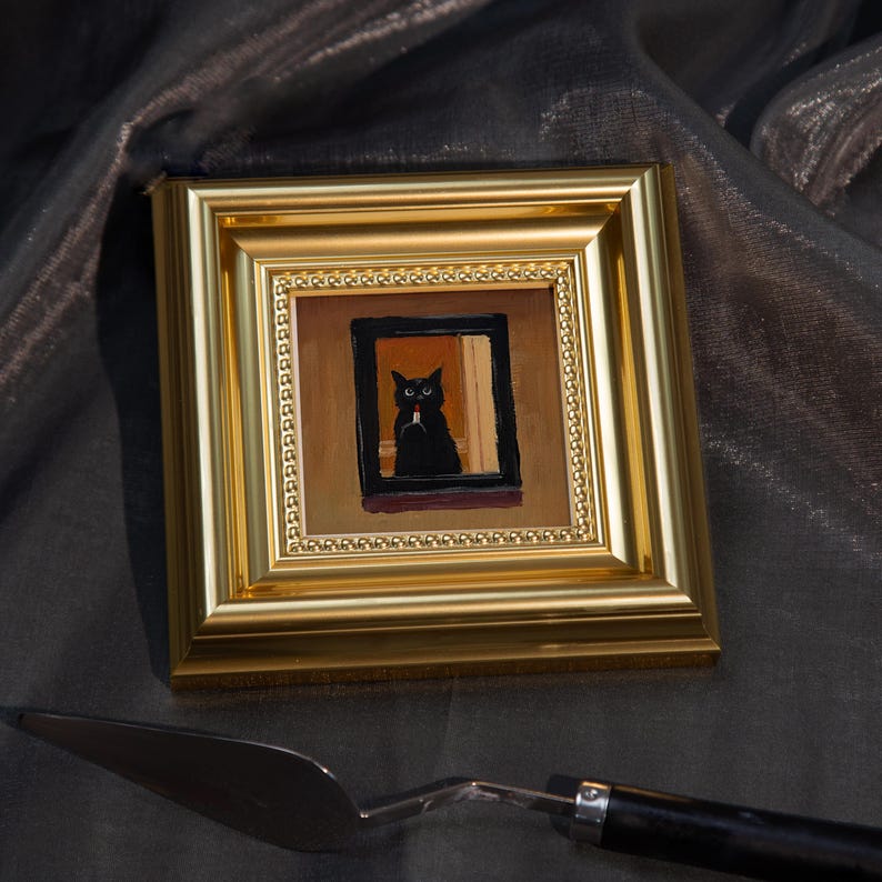 May include: A small, square painting in a gold frame depicts a black cat in a window. The cat is the focal point, with a white object in its mouth. The painting is set against a dark, textured background. A silver palette knife rests below.