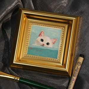 May include: A square, gold-framed painting of a white cat with pink ears and blue eyes, emerging from turquoise water. The painting is displayed on a gray surface, with a paintbrush and pen in the foreground.