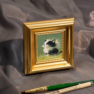 May include: A small framed painting of a black and white kitten with a gold frame. The painting has a green background and the kitten is the focal point. A green and gold paintbrush is in the foreground.