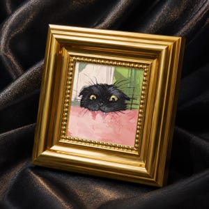 Könnte beinhalten: Ein gerahmtes Kunstwerk mit einer schwarzen Katze mit gelben Augen, die über eine rosa Oberfläche blickt. Das Fell der Katze ist detailliert, und das Kunstwerk ist in einem goldfarbenen Rahmen mit einem Perlenrand gefasst. Der Hintergrund enthält grüne und weiße Elemente.