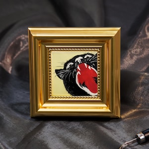 Könnte beinhalten: Ein gerahmtes Kunstwerk mit einem schwarzen Panther mit rotem offenem Maul und weißen Zähnen. Das Kunstwerk ist vor einem hellgelben Hintergrund platziert und in einem goldfarbenen Rahmen gerahmt. Der Rahmen hat einen Perlenrand.