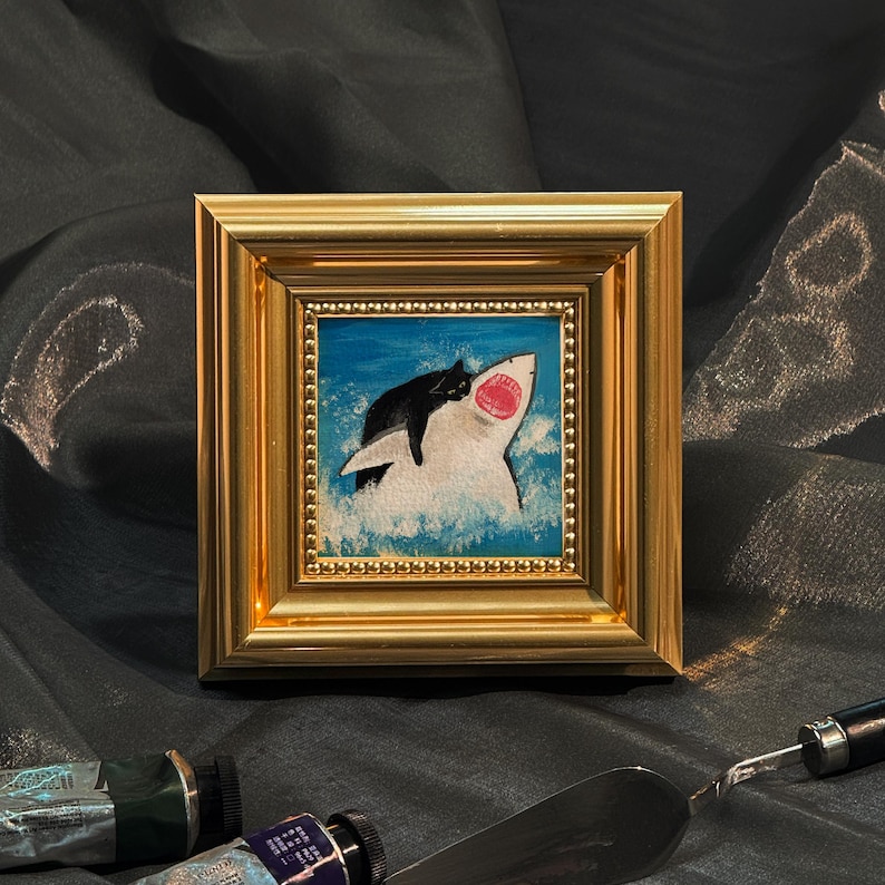 May include: A small, square painting in a gold frame depicts a black cat lounging on a white shark with an open mouth, set against a blue ocean background. The artwork is a whimsical, humorous piece, perfect for a home decor item.