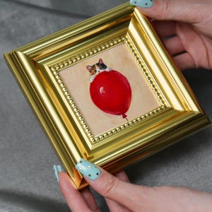 May include: A small, framed painting featuring a calico cat clinging to a large, red balloon. The artwork is set within a gold-colored frame with a beaded inner border. The painting has a whimsical, playful theme.
