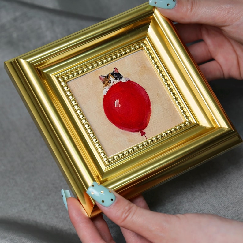 May include: A small, framed painting featuring a calico cat clinging to a large, red balloon. The artwork is set within a gold-colored frame with a beaded inner border. The painting has a whimsical, playful theme.