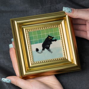 May include: A framed artwork featuring a black cat in a bathtub. The cat is depicted with yellow eyes and is standing in water. The frame is gold-colored and square. The background shows green tiles.