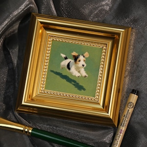 Può includere: Un dipinto incorniciato in oro di un cucciolo bianco, nero e marrone a mezz'aria su uno sfondo verde. Un pennello con manico verde e una penna nera sono in primo piano. Il dipinto è di forma quadrata.